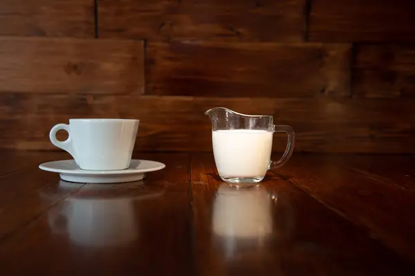 Latte in brocca di latte di vetro con tazza bianca e riflessione da un tavolo di legno — Foto Stock