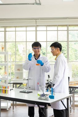 Bilim, Kimya, Teknoloji, Biyoloji ve Laboratuar Konsepti - Asyalı kıdemsiz bilim adamı deney sonuçlarını cam deney şişesinde gösterirken, Asyalı kıdemli bilim adamı bir öneri sunuyor.