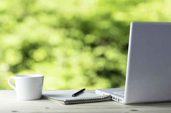 有白色笔记本电脑 笔记本电脑和咖啡在木制桌子上的工作场所 背景在一个模糊的自然绿色花园中 — 图库照片