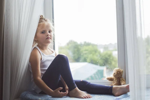 Chica triste sentado en el alféizar de la ventana y mirando —  Fotos de Stock