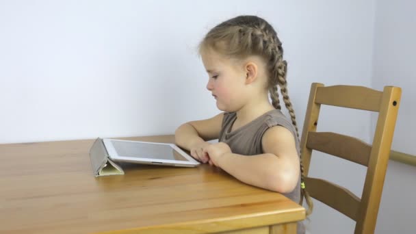 Niña usa la tableta mientras está sentada en la mesa — Vídeos de Stock