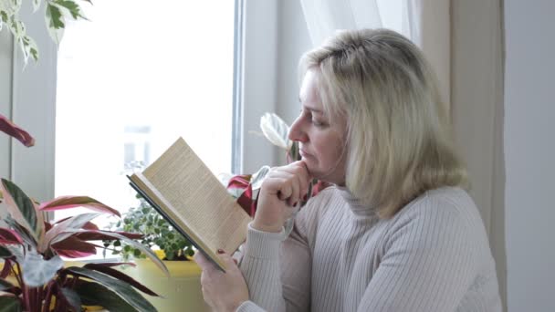 Mujer adulta se sienta junto a la ventana y lee un libro — Vídeo de stock