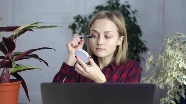 Flicka målar ögonfransar och fungerar bakom en laptop — Stockvideo