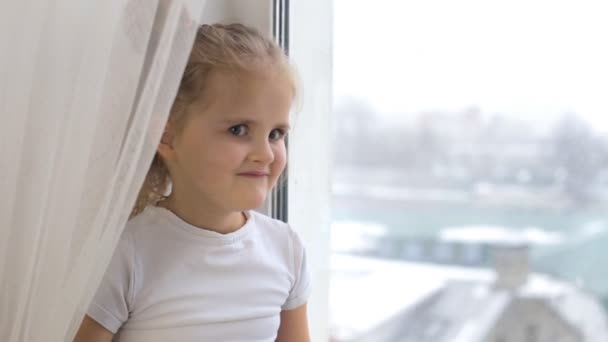 La niña está sentada junto a la ventana y mirando a la cámara con una sonrisa — Vídeos de Stock