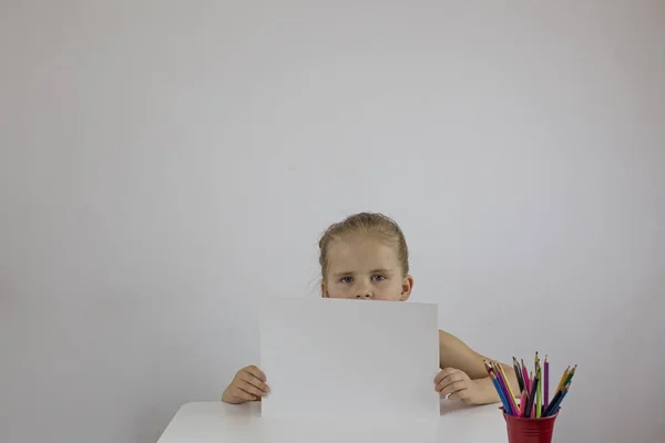 Dziewczyna trzyma białą kartkę, siedząc przy biurku na białej ścianie — Zdjęcie stockowe