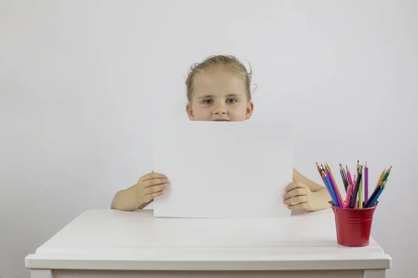 Little schoolgirl sitting at her desk t holding a white sheet of paper — Stock Photo, Image