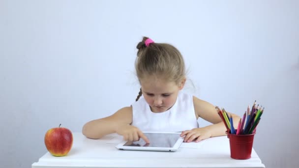 Kislány ül az íróasztalon, és végrehajtja a feladatot a tabletta — Stock videók