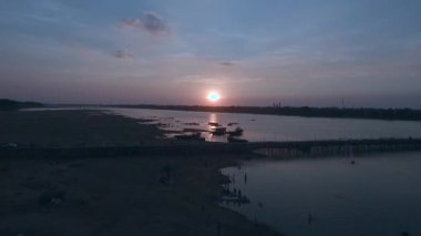 Hava: nehir kıyısında günbatımı, çevresinde Yüzme insan siluetleri ile bambu köprüden üzerinde sinek demirleyen tekneleri