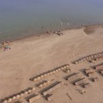 Fly back revealing straw huts and wooden bungalows spread over sand beach. People swimming along the beach as backdrop