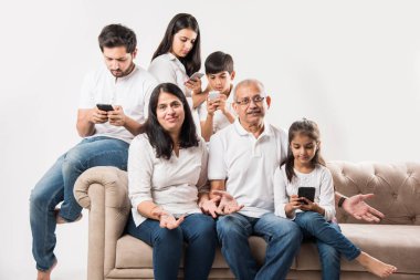 Yaşlı yetişkinler sıkılırken ve genç üyeler akıllı telefondan meşgulken Hintli aile koltukta oturuyor.