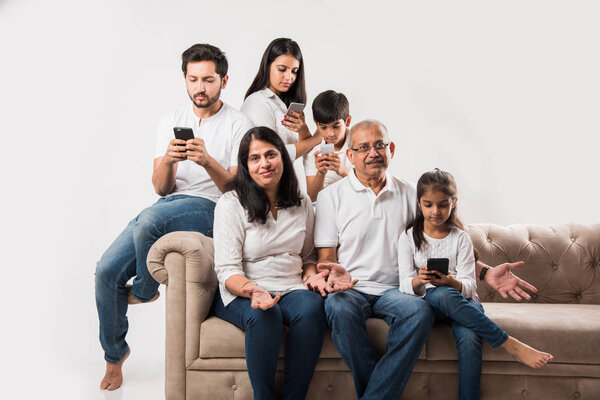 Индийская семья сидит на диване, в то время как пожилые люди скучают и молодые члены заняты на смартфоне