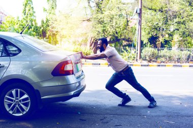 Hintli sakallı adam bütün gücüyle bozuk arabayı yola itiyor.