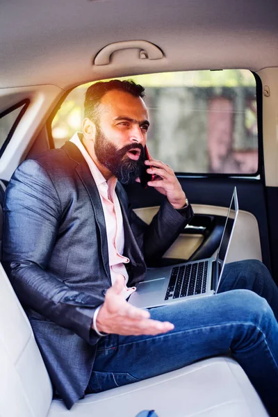 Indian/asian young businessman using laptop computer and phone inside the car, selective focus