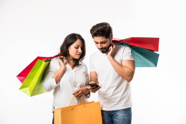 Hintli genç çift alışveriş çantaları ve akıllı telefon veya cep telefonu, selfie veya mağaza bulma tıklayarak, beyaz arka plan üzerinde izole ayakta. seçici odaklama