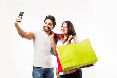 Hintli genç çift alışveriş çantaları ve akıllı telefon veya cep telefonu, selfie veya mağaza bulma tıklayarak, beyaz arka plan üzerinde izole ayakta. seçici odaklama