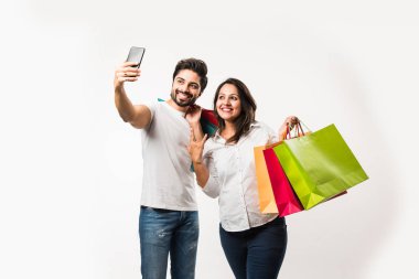 Hintli genç çift alışveriş çantaları ve akıllı telefon veya cep telefonu, selfie veya mağaza bulma tıklayarak, beyaz arka plan üzerinde izole ayakta. seçici odaklama