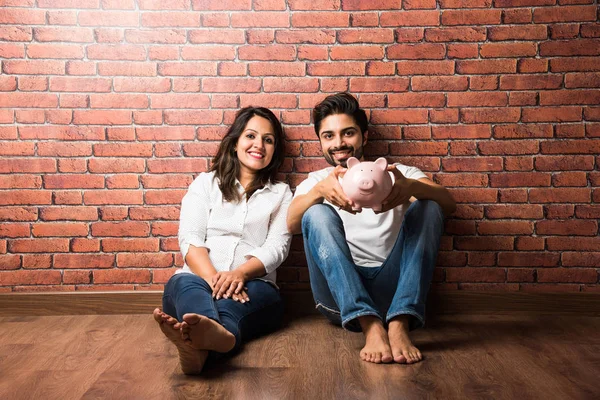 Indian couple with piggy bank sitting in front of brick background
