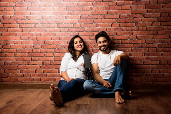 Indian smart couple listen to music on headphones, sitting against exposed brick wall inside home