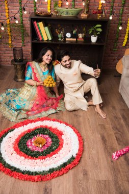 Hintli çift Diwali veya Onam Festivali'nde çiçek Rangoli yapma, selfie alarak veya tatlılar tutarak