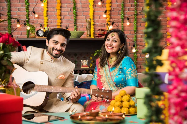 印度丈夫在迪瓦利节或周年纪念日为妻子弹吉他 周围有糖果 迪雅和礼品盒 而坐在室内的坐式坐 — 图库照片