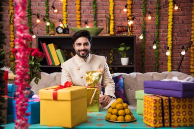 Geleneksel kıyafet giyerken festival gününde tablet dokunmatik bilgisayar kullanan Hintli adam. aydınlatma bokeh ile hediyeler ve tatlılar ile kanepede oturan