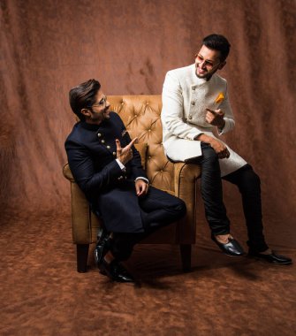 İki Hintli erkek etnik ya da geleneksel kıyafetler giyer, Sherwani ya da Kurta pijaması olan erkek modeller, kahverengi grunge arka planına karşı oturur ve kanepede poz verirler, seçici bir odak noktası vardır.