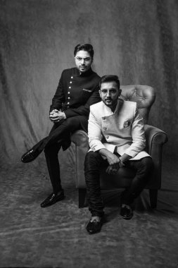 İki Hintli erkek etnik ya da geleneksel kıyafetler giyer, Sherwani ya da Kurta pijaması olan erkek modeller, kahverengi grunge arka planına karşı oturur ve kanepede poz verirler, seçici bir odak noktası vardır.