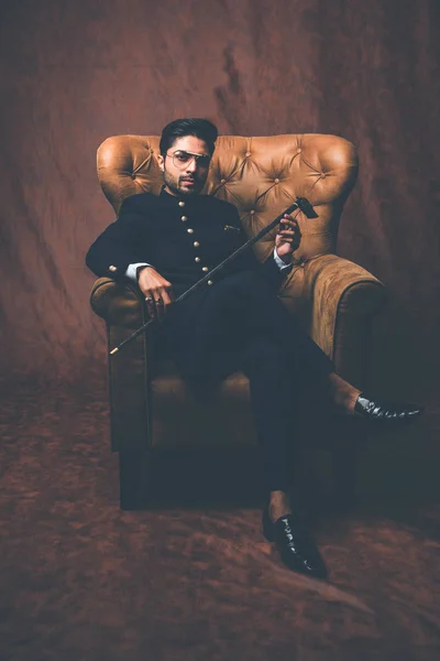 Indian bridegroom wears ethnic or traditional cloths,  Male fashion model with dark blue sherwani, Sitting on Wing Chair / sofa over brown grunge background, selective focus