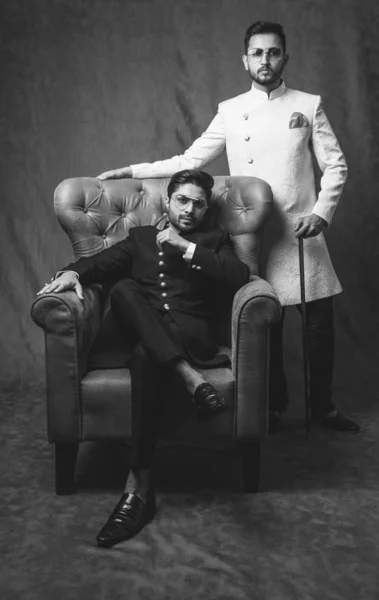 two Indian men wears ethnic or traditional cloths,  Male fashion models with sherwani or Kurta Pyjama, sitting and posing on wing chair or sofa against brown grunge background, selective focus