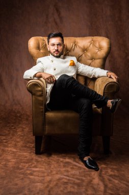 Hintli damat etnik ya da geleneksel giysiler giyer, koyu mavi sherwani 'li erkek model, kahverengi grunge arka planında Kanat Sandalyesi / kanepede oturan, seçici odaklı