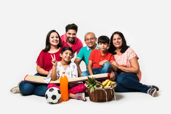 Indian Family enjoying Picnic indoors - Multi generation of asian family sitting over over white background  with fruit basket, mat, football and drinks. selective focus