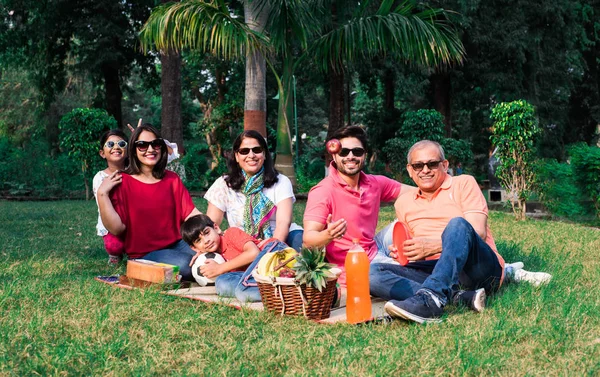 Indian Family enjoying Picnic - Multi generation of asian family sitting over lawn or green grass in park with fruit basket, mat and drinks. selective focus