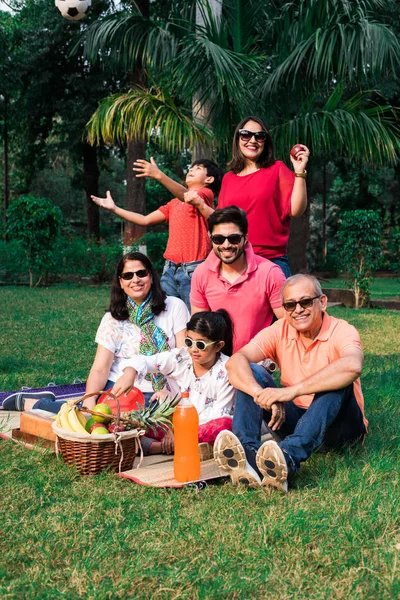 Indian Family enjoying Picnic - Multi generation of asian family sitting over lawn or green grass in park with fruit basket, mat and drinks. selective focus