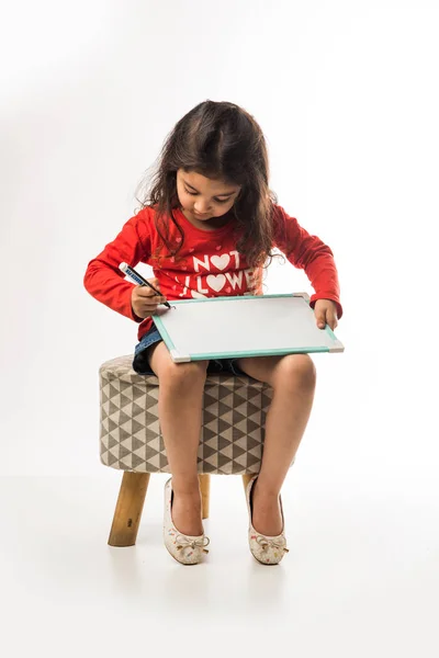 small Indian girl writing on slate with marker pen, while sitting on stool over white background