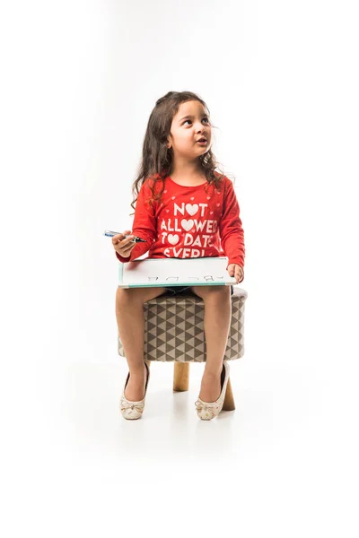 small Indian girl writing on slate with marker pen, while sitting on stool over white background