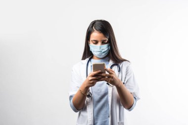 Hintli kadın doktor akıllı telefondan konuşuyor. Heyecanlı, koronavirüs salgınında tıbbi maske takıyor.