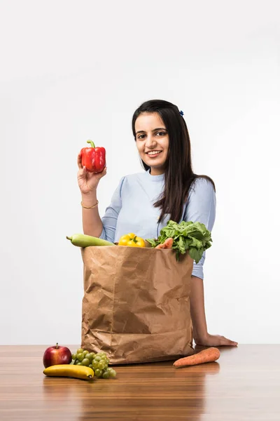 Close up portrait of a happy pretty Indian girl holding bag with vegetables and fruits standing isolated over white background