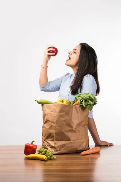 Close up portrait of a happy pretty Indian girl holding bag with vegetables and fruits standing isolated over white background