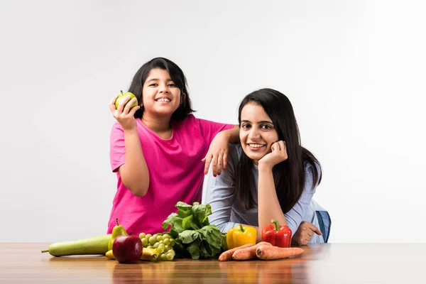 Beautiful Indian / Asian young Mother and Daughter posing or playing at table full of fruits and vegetables