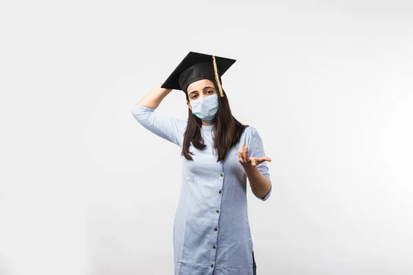 Corona Pandemic and confusion over university exams in India - Pretty Indian Female student with confused expressions wearing medical face mask and graduation hat