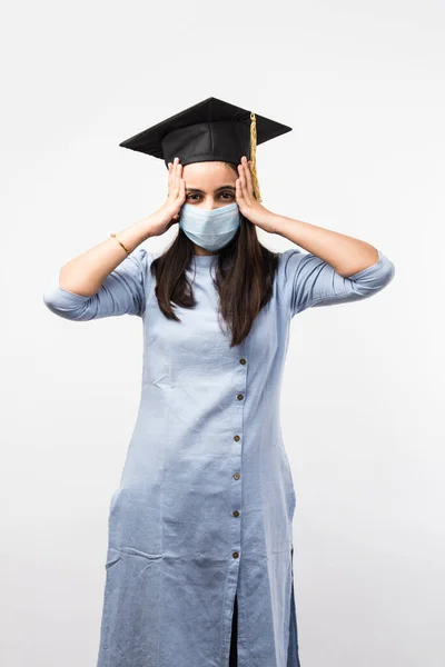 Corona Pandemic and confusion over university exams in India - Pretty Indian Female student with confused expressions wearing medical face mask and graduation hat