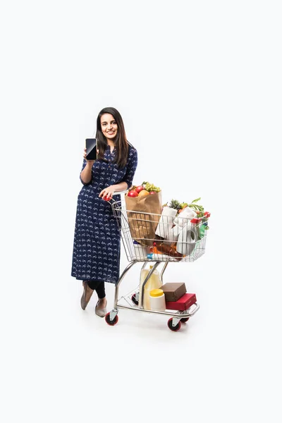 Indian young woman with shopping cart or trolly full of grocery, vegetables and fruits.  Isolated Full length photo over white background