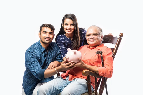 Indian old man or father holding piggy bank with young son and daughter, sitting over rocking chair