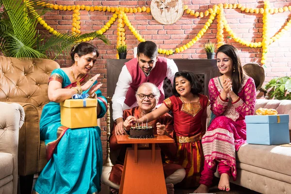 Indian old man with Family celebrating birthday by blowing candles on cake while wearing ethnic wear