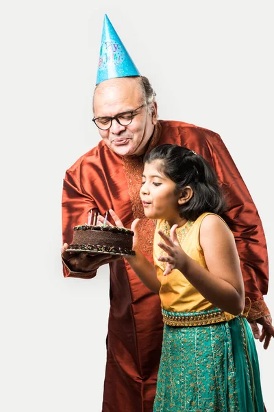 Indian senior or old man with granddaughter celebrating birthday by blowing candles on cake while wearing ethnic wear, standing isolated against white background