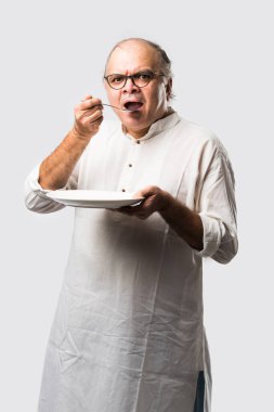 Yaşlı ya da yaşlı Hintli bir adam kaşıkla çatal kullanarak boş beyaz tabak ya da kâseden yemek yiyor. Beyaz arka planda farklı ifadelerle tek başına duruyor.