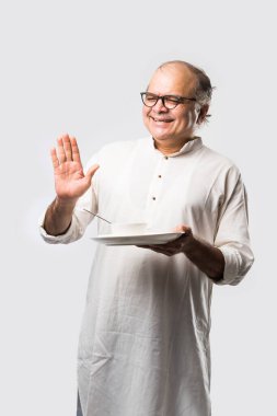 Yaşlı ya da yaşlı Hintli bir adam kaşıkla çatal kullanarak boş beyaz tabak ya da kâseden yemek yiyor. Beyaz arka planda farklı ifadelerle tek başına duruyor.