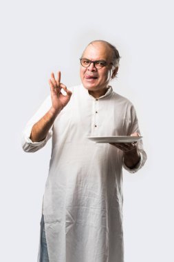 Yaşlı ya da yaşlı Hintli bir adam kaşıkla çatal kullanarak boş beyaz tabak ya da kâseden yemek yiyor. Beyaz arka planda farklı ifadelerle tek başına duruyor.