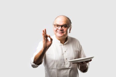 Yaşlı ya da yaşlı Hintli bir adam kaşıkla çatal kullanarak boş beyaz tabak ya da kâseden yemek yiyor. Beyaz arka planda farklı ifadelerle tek başına duruyor.