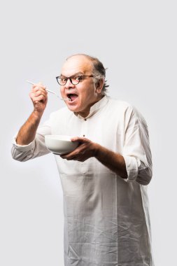 Yaşlı ya da yaşlı Hintli bir adam kaşıkla çatal kullanarak boş beyaz tabak ya da kâseden yemek yiyor. Beyaz arka planda farklı ifadelerle tek başına duruyor.